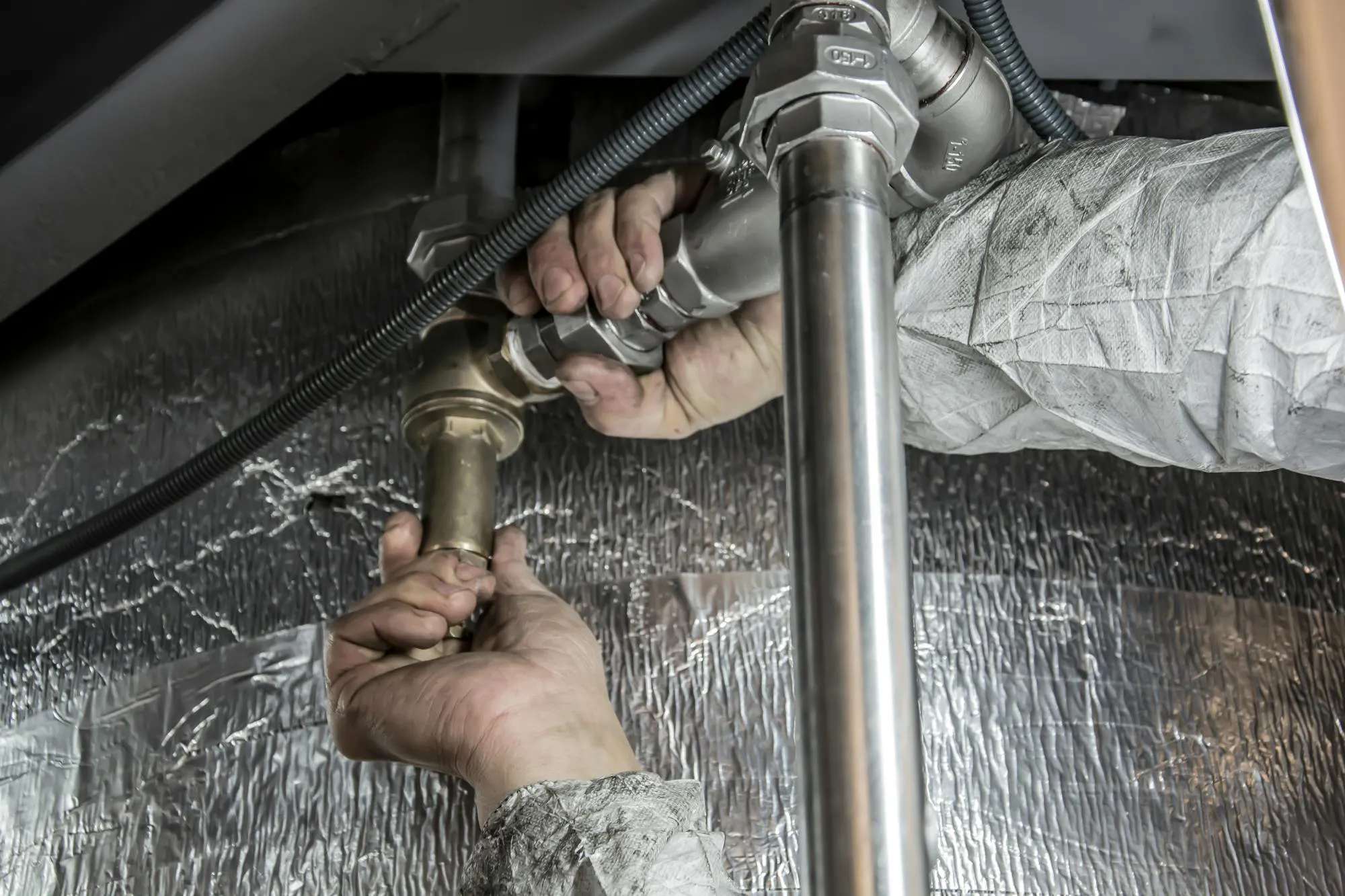 Hands fitting a brass and steel pipework joint under a kitchen unit in Chelmsford.